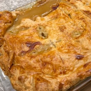Golden brown pastry covers a homemade chicken pie in a rectangular dish, with gravy oozing out.