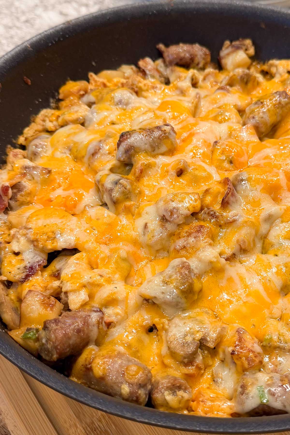 A skillet filled with a cheesy, golden-brown casserole. Chunks of sausage, bacon, potatoes, and mushrooms.