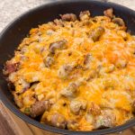 A skillet filled with a cheesy, golden-brown casserole. Chunks of sausage, bacon, potatoes, and mushrooms.