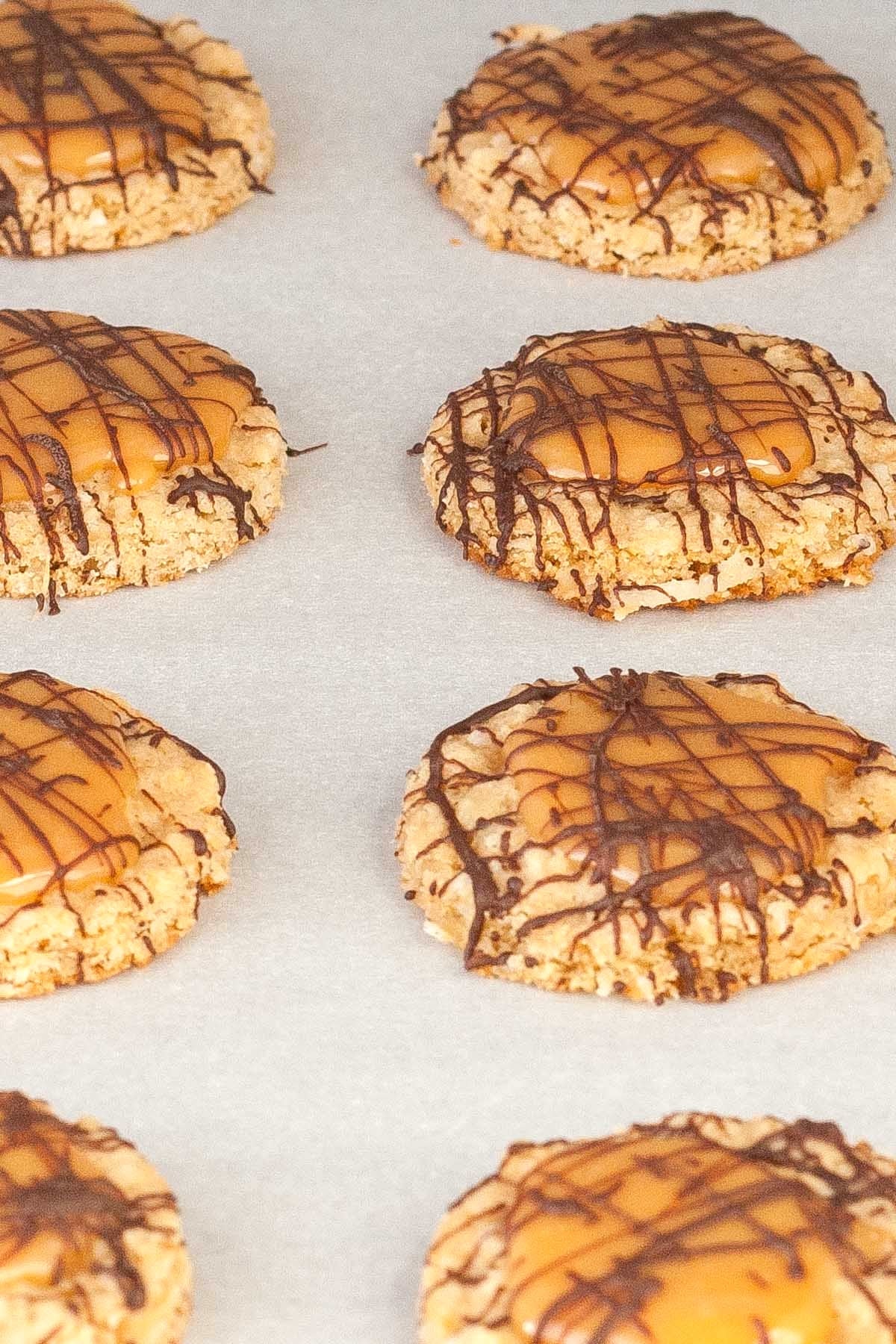 Oatmeal cookies topped with caramel and drizzled with chocolate rest on a parchment-lined baking sheet.