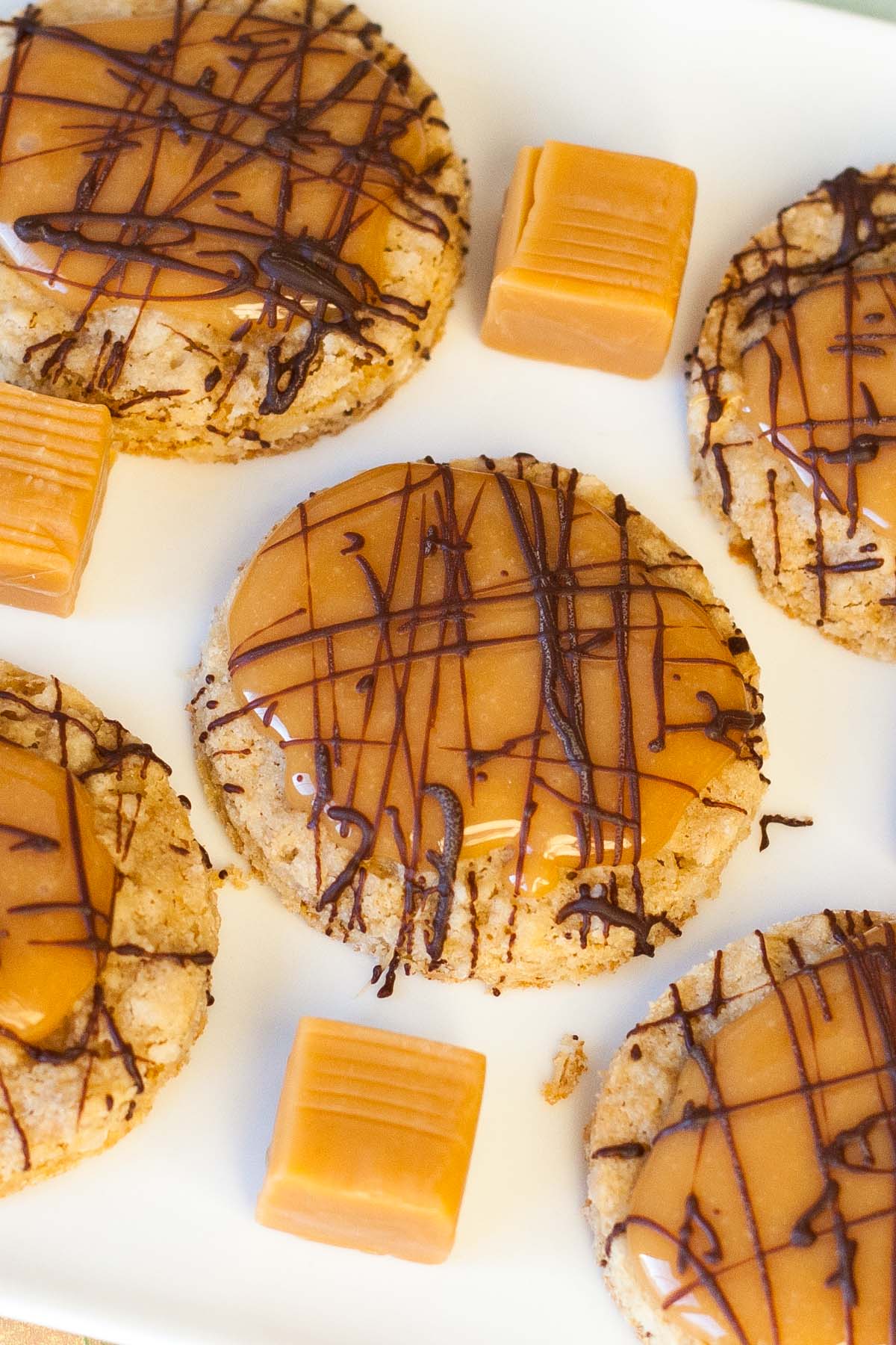 Oatmeal cookies topped with caramel and drizzled with chocolate on a white plate. Caramel squares are placed between the cookies.