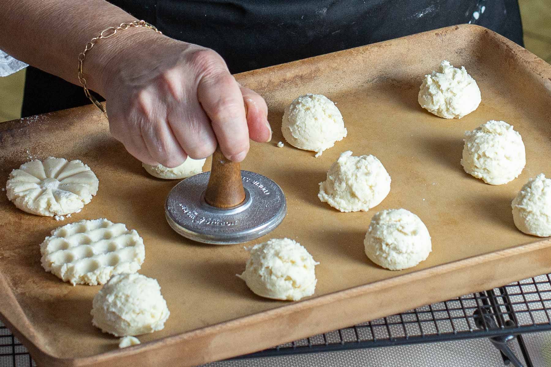 A hand presses a wooden stamp onto cookie dough balls on a baking sheet, creating decorative patterns.