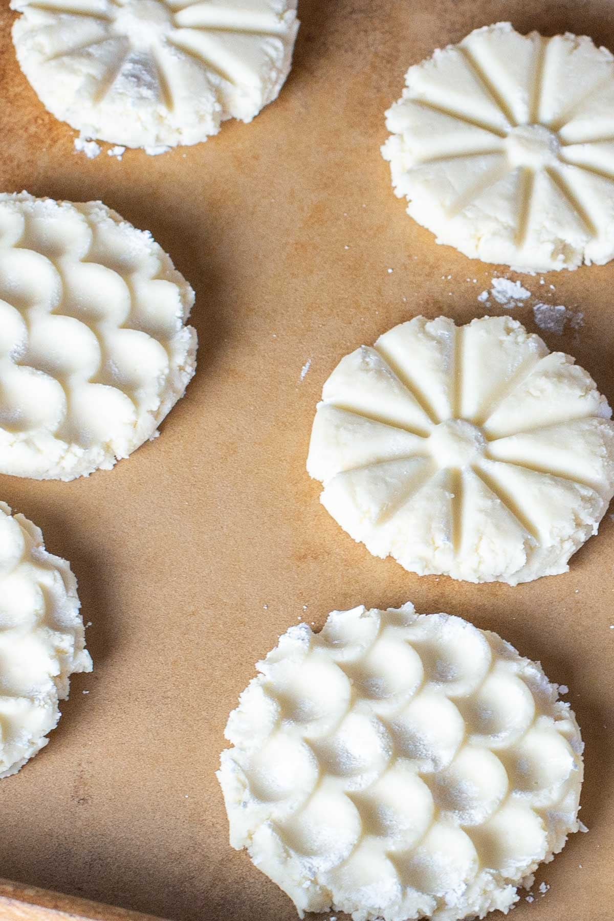 Six unbaked shortbread cookies with intricate patterns rest on a parchment-lined baking sheet.