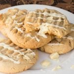 A plate of white chocolate pudding cookies drizzled with white chocolate. One cookie has a bite taken, revealing a soft texture.