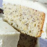 A slice of banana cake with white frosting is lifted from a cake decorated with chocolate slivers on a crystal platter.