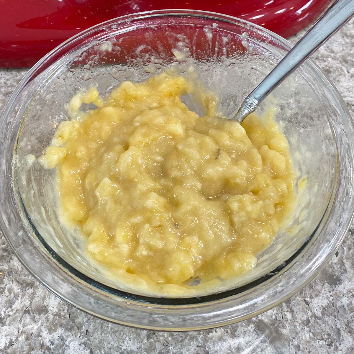 A glass bowl filled with mashed bananas and a spoon, set on a grey speckled countertop.