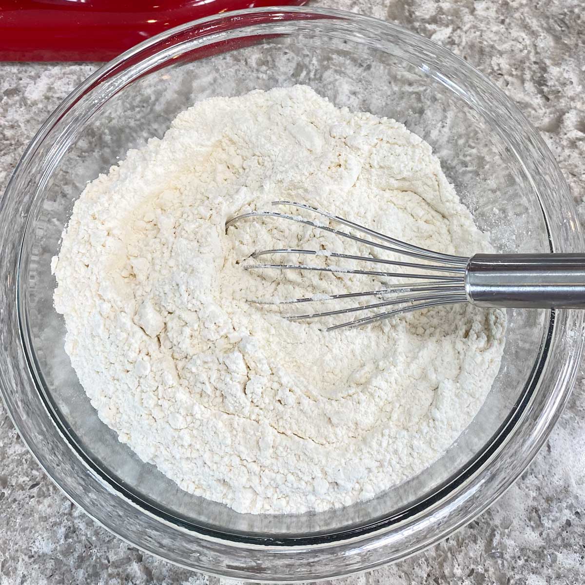 A glass bowl of flour is being whisked on a textured gray countertop.