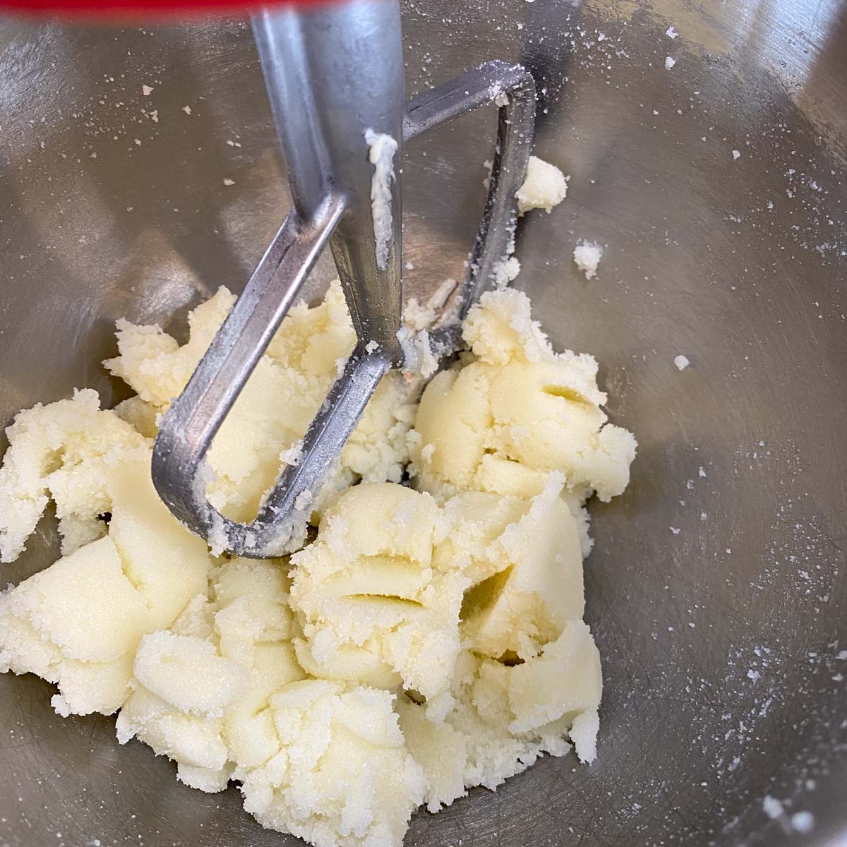 A close-up of creamed butter and sugar in a metal mixing bowl. A metal paddle attachment is visible above the mixture.