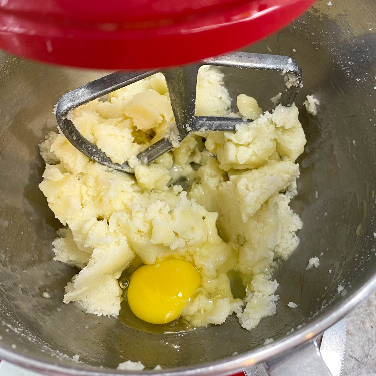 A close-up of creamed butter and sugar in a mixing bowl with a single egg yolk. A metal paddle attachment is visible above the mixture.