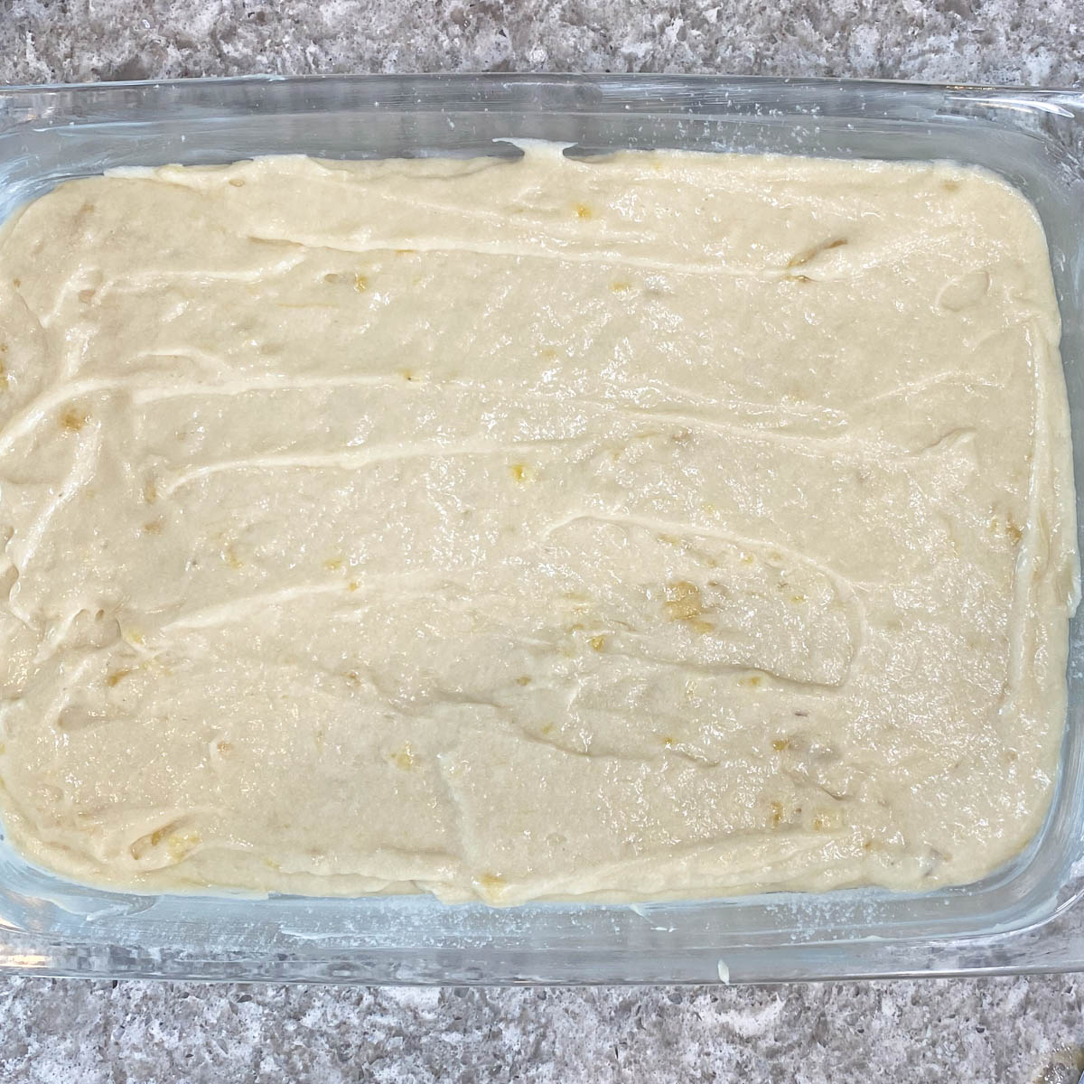 Smooth, creamy, banana cake batter evenly spread in a rectangular glass baking dish on a speckled gray countertop, ready for baking.