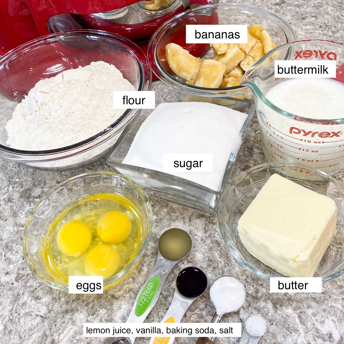 Labeled baking ingredients on a countertop include flour, sugar, eggs, butter, buttermilk, and bananas. Measuring spoons with lemon juice, vanilla, baking soda, and salt lie nearby.