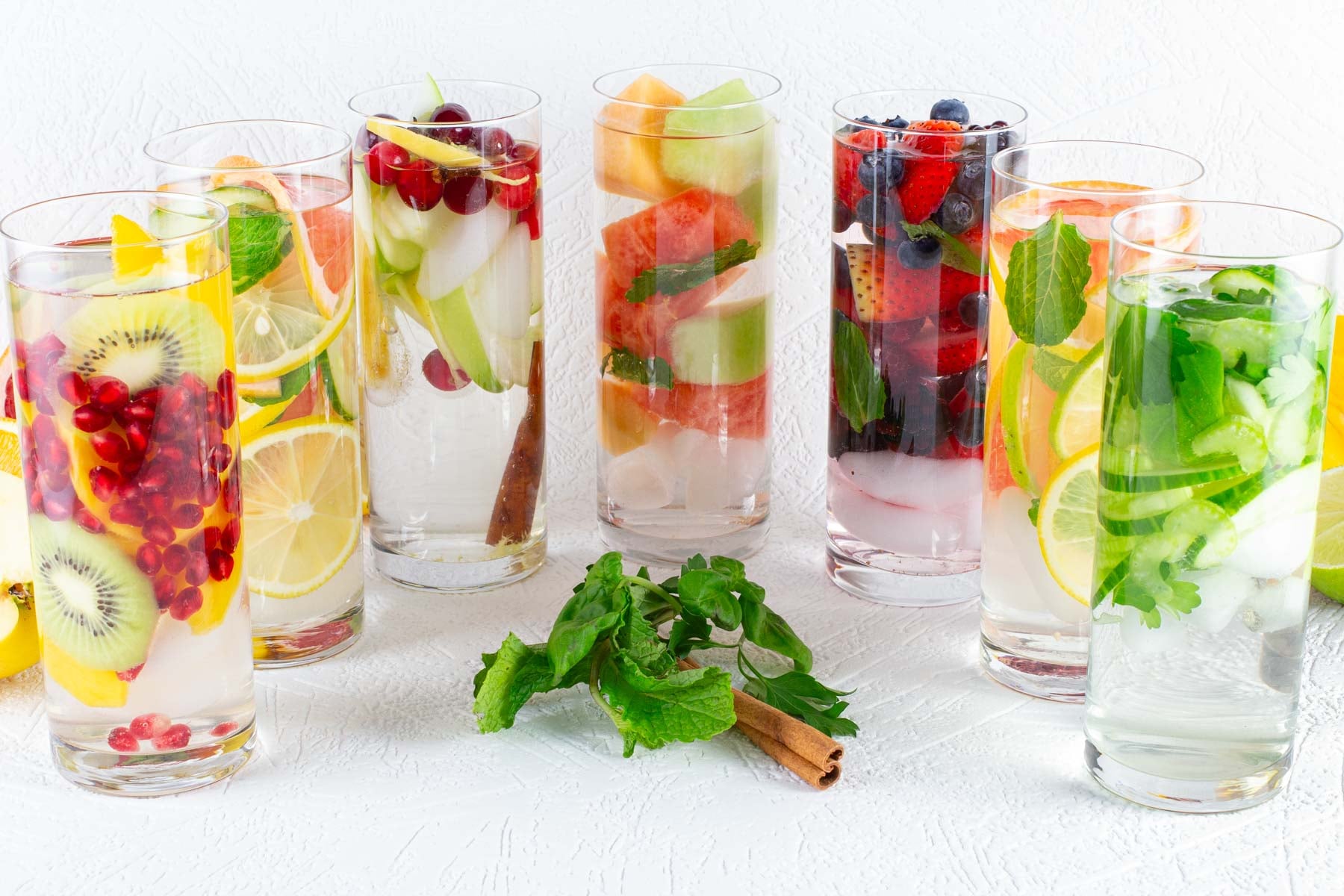 Colorful infused water glasses lined up, each filled with assorted fruits like kiwi, berries, citrus, and herbs like mint.