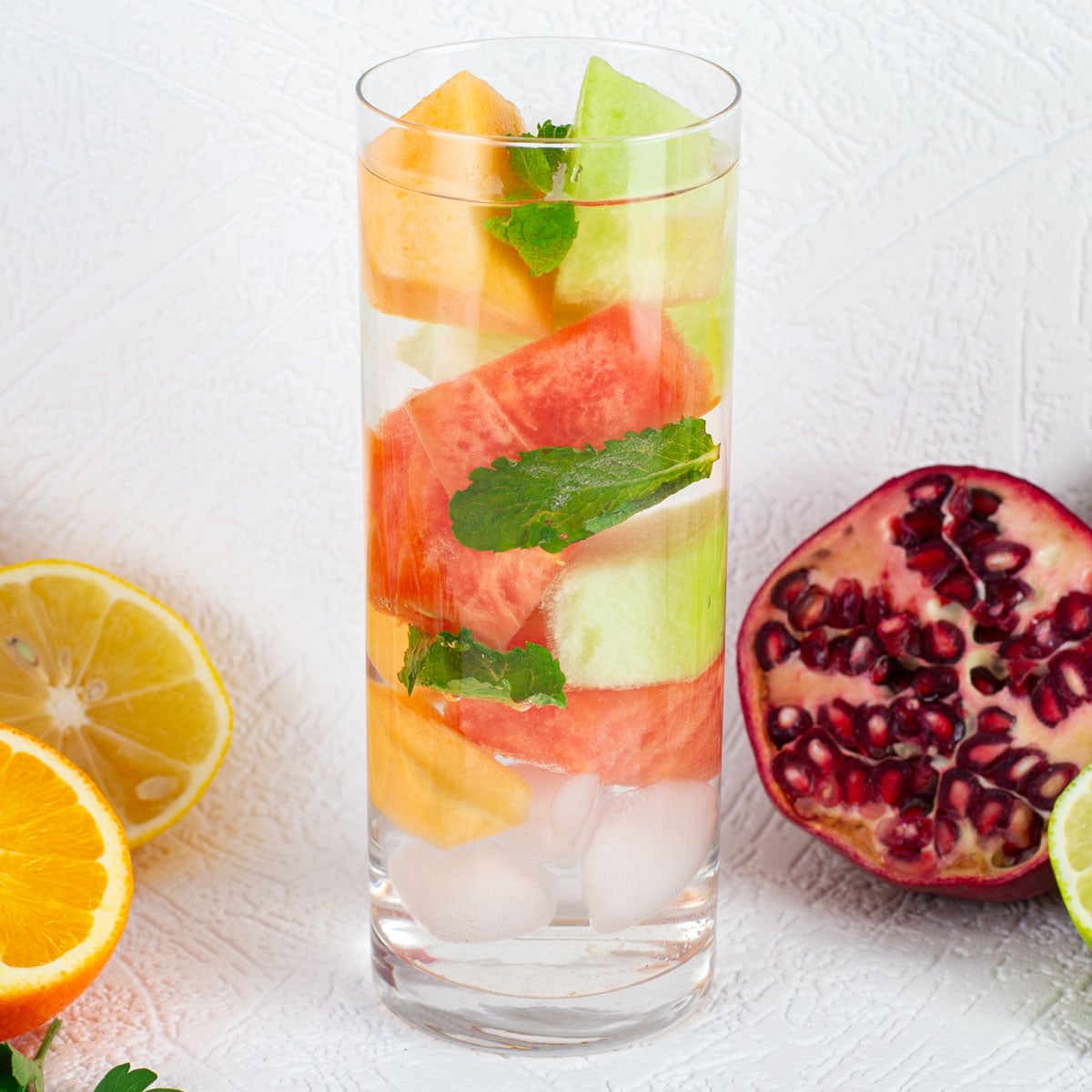 A tall glass filled with ice, colorful fruit slices, and mint leaves. Surrounded by sliced lemon, orange, and a halved pomegranate.