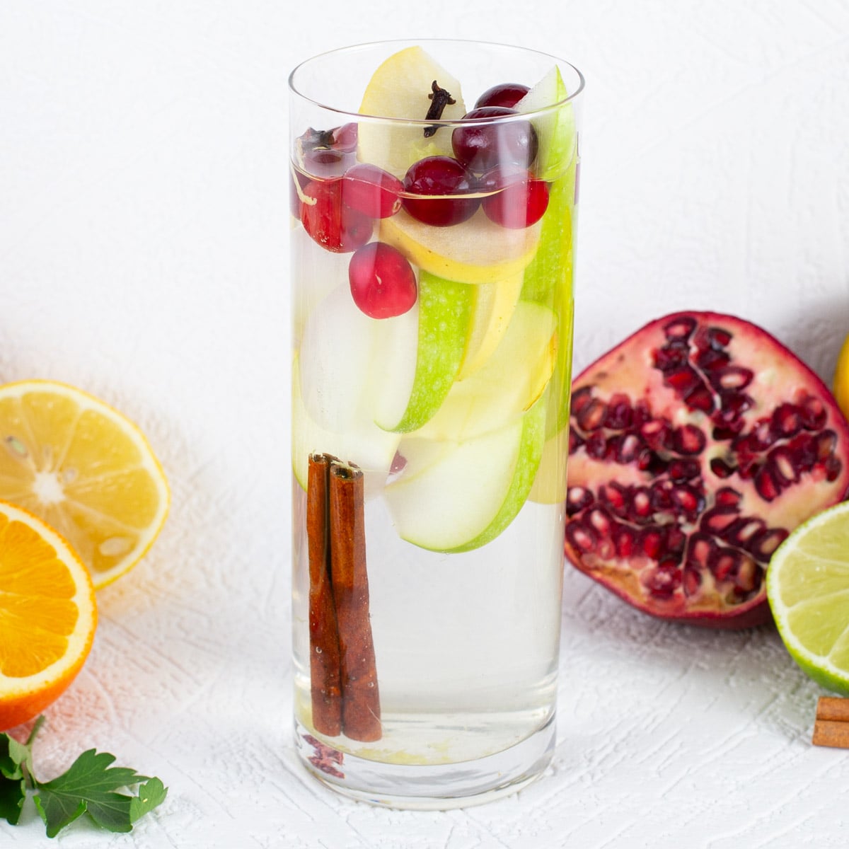 A tall glass of water infused with apple slices, cranberries, and cinnamon sticks. Surrounded by lemon, orange, and pomegranate on a textured white surface.