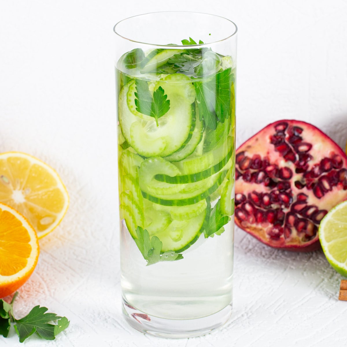 A tall glass filled with water, cucumber slices, and parsley, surrounded by half-sliced lemon, orange, pomegranate, and lime on white background.