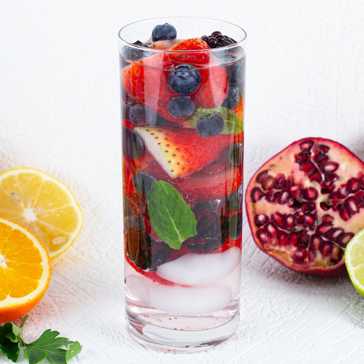 A tall glass of fruit-infused water with strawberries, blueberries, mint, and ice. Surrounded by sliced citrus fruits and a halved pomegranate.