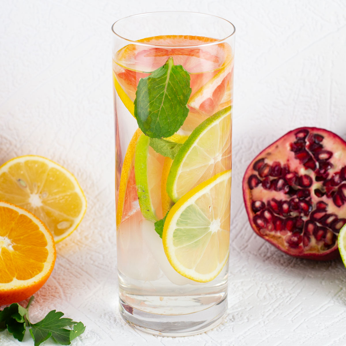 A tall glass of infused water with sliced citrus fruits and mint. Surrounding fresh orange, lemon, and pomegranate on a white background.