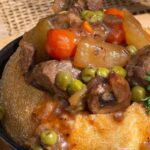 A savory bowl with beef stew, carrots, peas, and mushrooms in a baked bread bowl, garnished with fresh parsley.