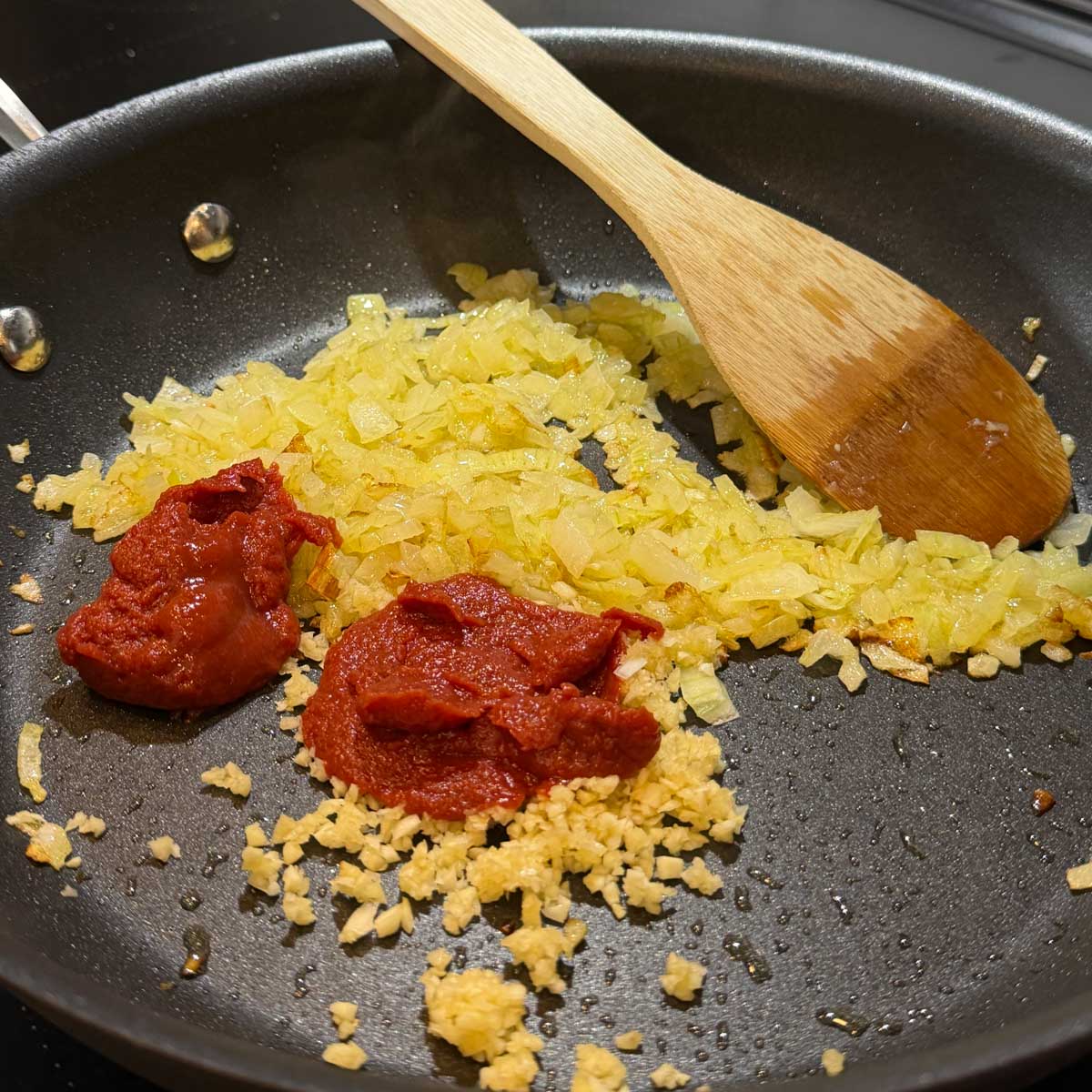 Chopped onions, tomato paste, and minced garlic are sizzling in a black skillet with a wooden spoon.