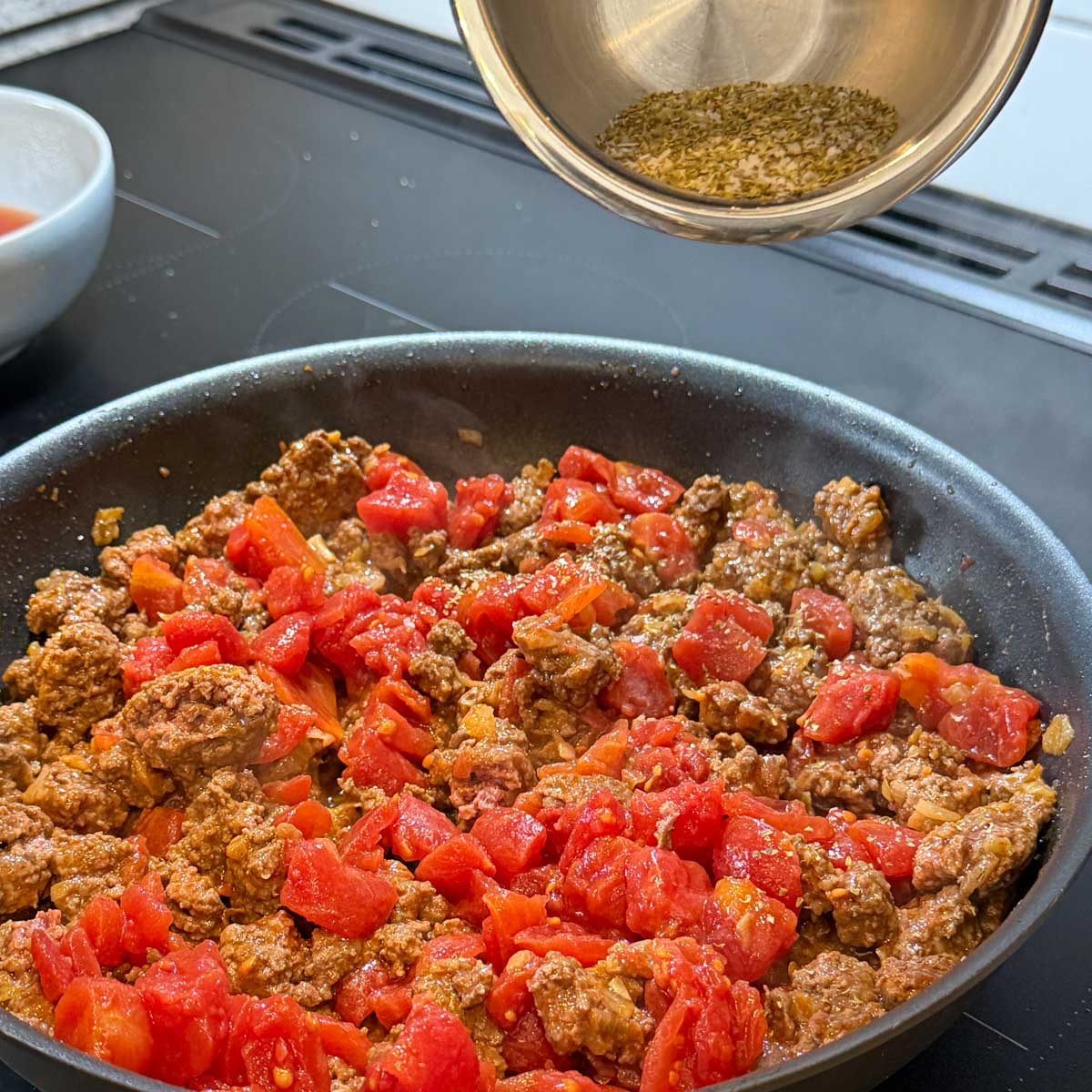 Ground beef and diced tomatoes cooking in a skillet, with dried spices being sprinkled from a metal bowl above.