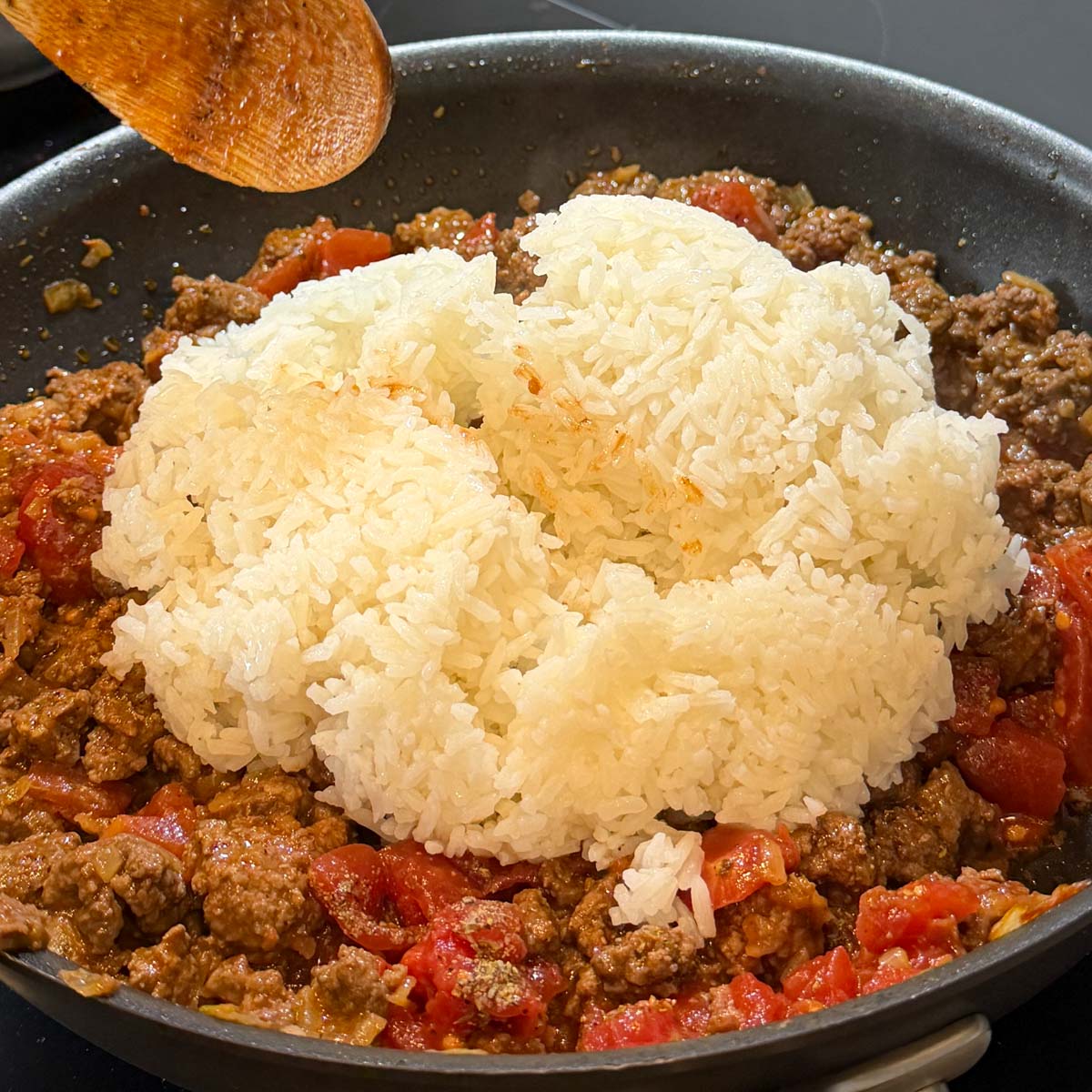 A skillet with cooked ground beef, tomatoes, and spices, topped with a mound of white rice. A wooden spoon hovers above, ready to stir.