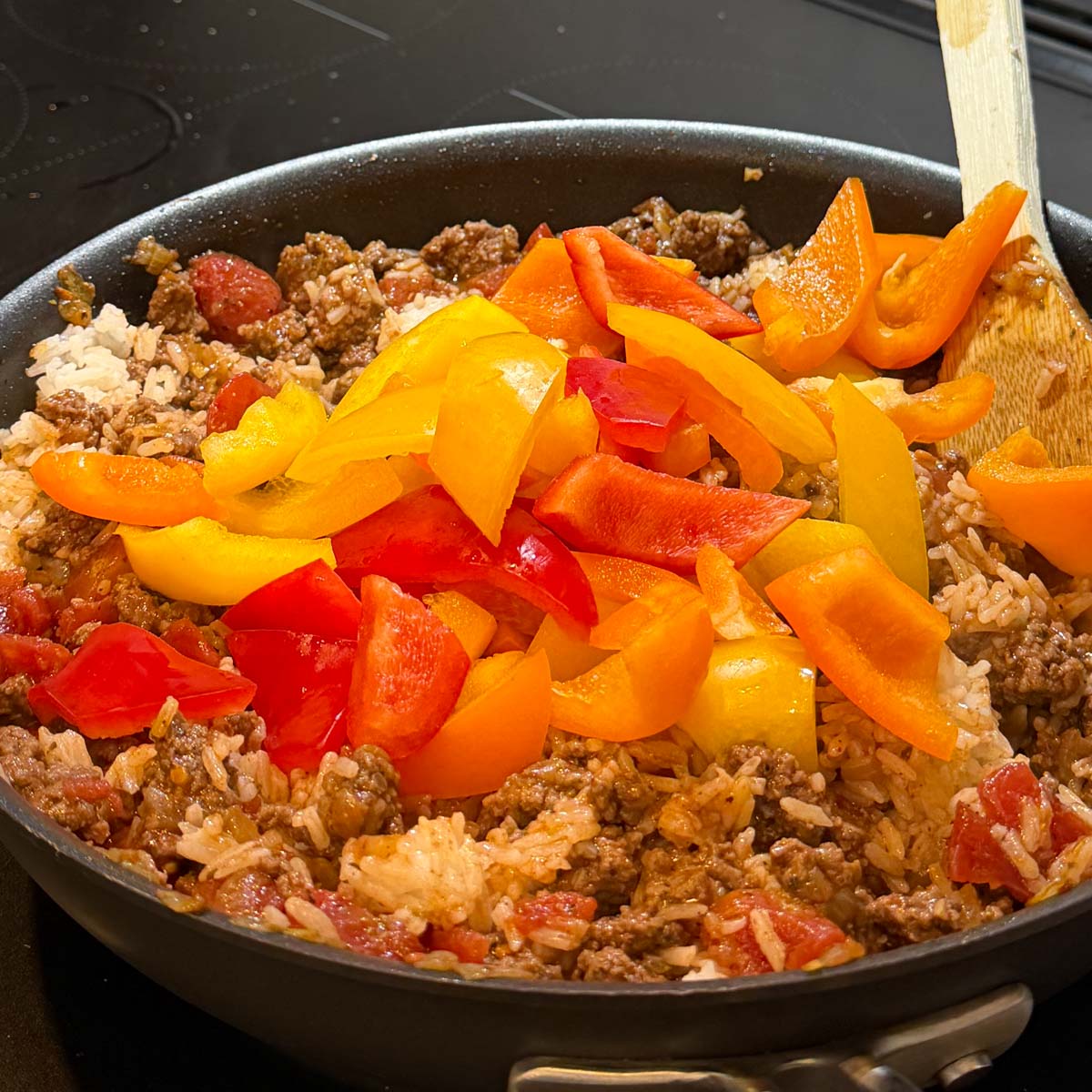 A skillet filled with saut&eacute;ed ground beef, white rice, diced tomatoes, topped with sliced red, orange, and yellow bell peppers, with a wooden spoon.