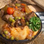 A savory bowl with beef stew, carrots, peas, and mushrooms in a baked bread bowl, garnished with fresh parsley.