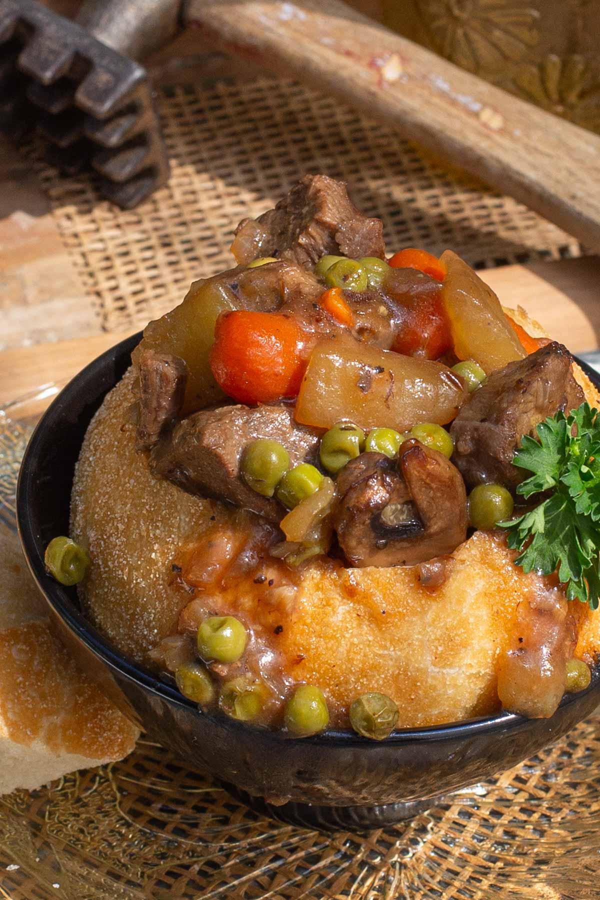 A savory bowl with beef stew, carrots, peas, and mushrooms in a baked bread bowl, garnished with fresh parsley. A wooden meat mallet is in the background.