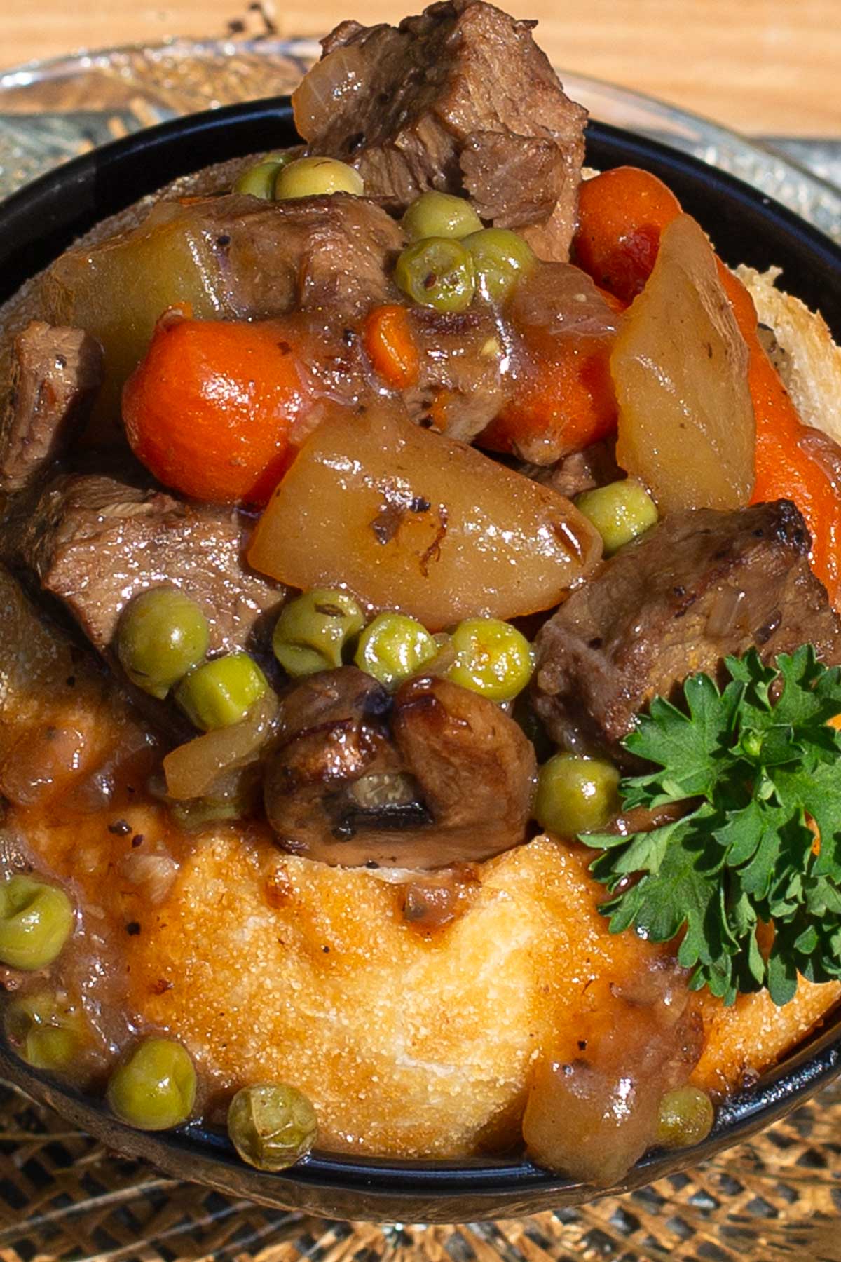 A savory bowl with beef stew, carrots, peas, and mushrooms in a baked bread bowl, garnished with fresh parsley.