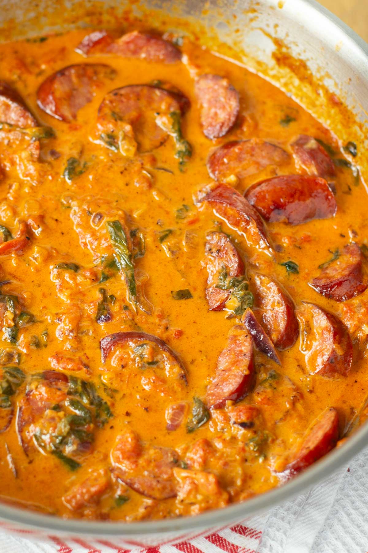 Creamy sauce in a stainless steel skillet with sliced sausage, spinach, and tomatoes.
