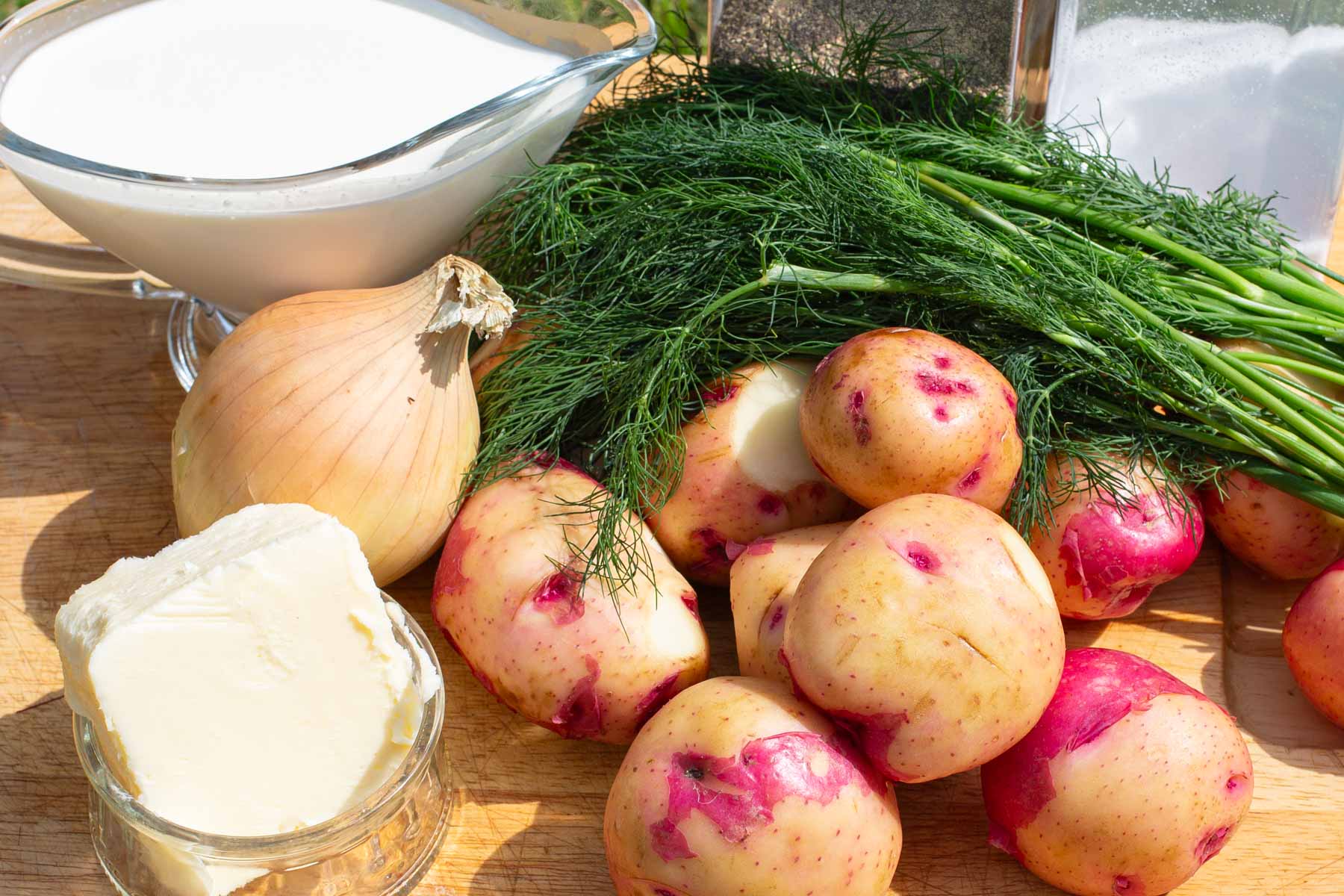 Fresh ingredients on a wooden surface: red-skinned potatoes, a whole onion, fresh dill, butter in a dish, and a bowl of cream.