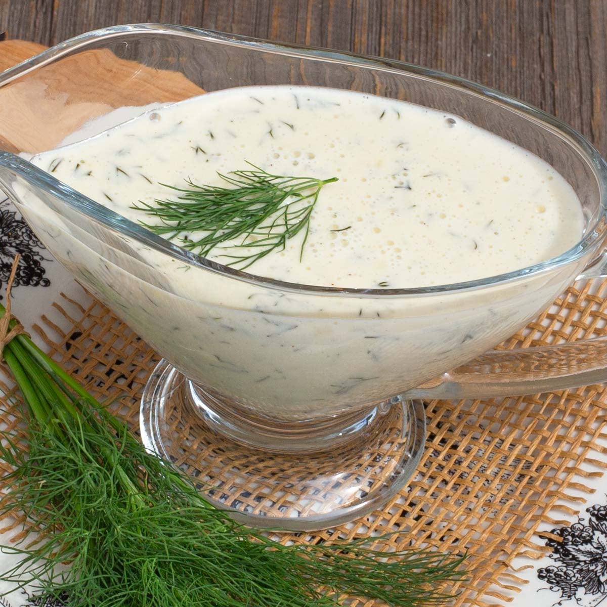 Glass gravy boat filled with creamy dill sauce, garnished with fresh dill. Set on a woven mat.