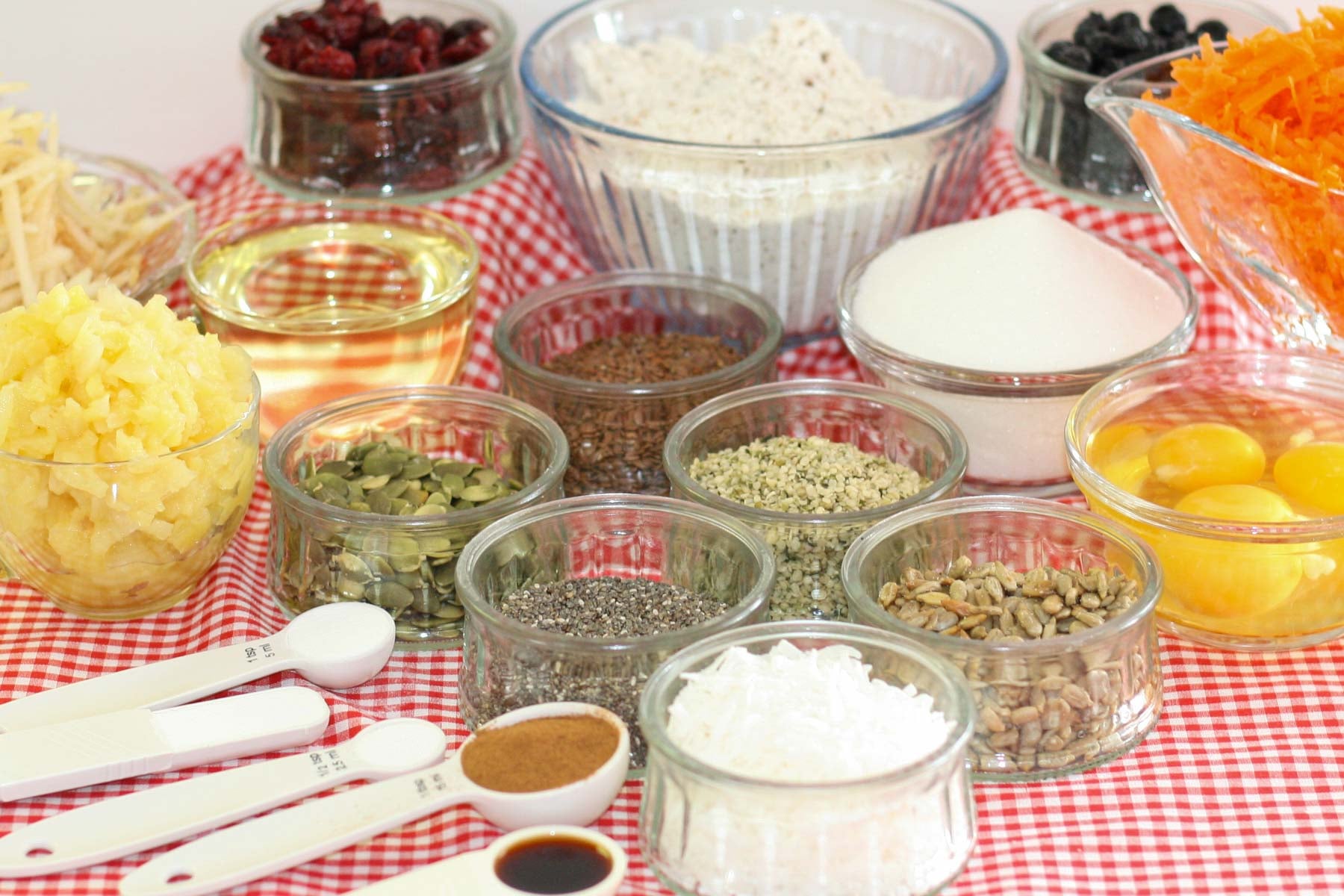 Assorted baking ingredients on a red-checkered cloth include flour, sugar, eggs, seeds, grated carrots, and spices, arranged in glass bowls.