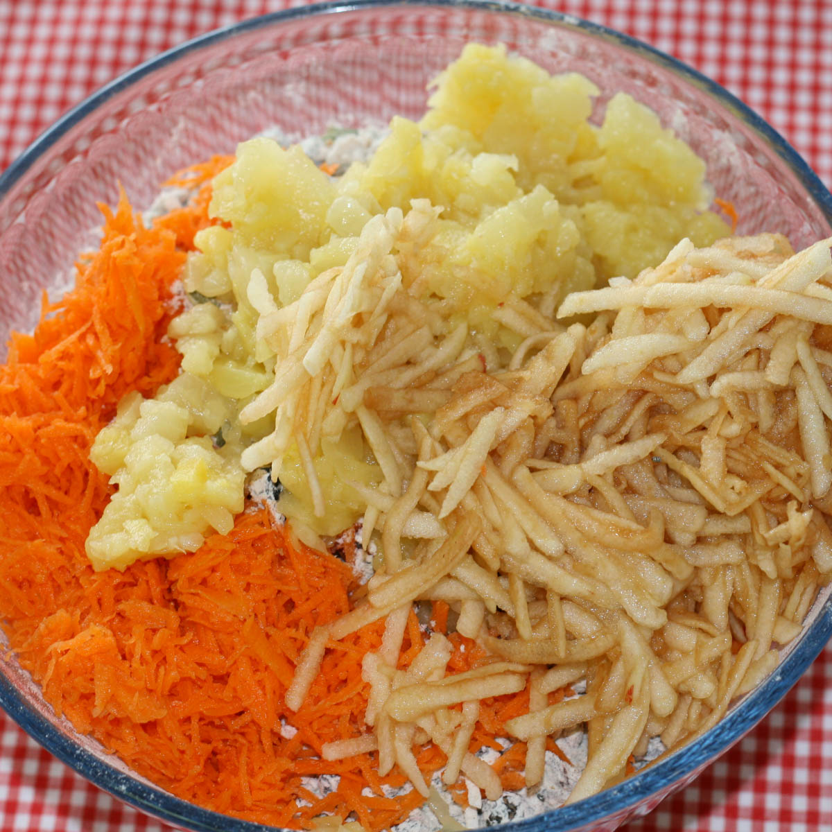 A glass bowl contains grated carrots, shredded apples, and crushed pineapple on a red and white checkered tablecloth.