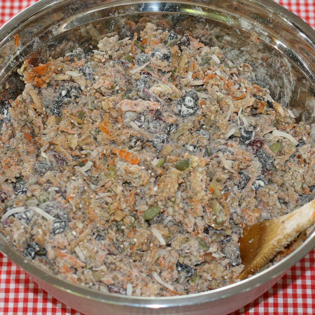 Stainless steel bowl filled with a chunky mixture of muffin batter, grated carrots, seeds, coconut, dried fruit, stirred with a wooden spoon.