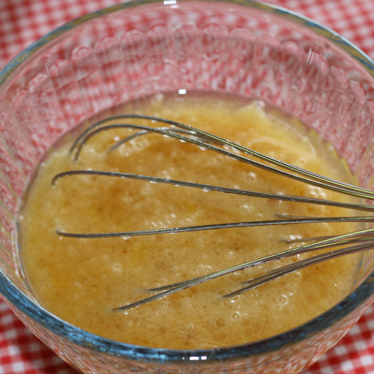 A glass bowl filled with lightly beaten eggs and a metal whisk, set on a red and white checkered surface.