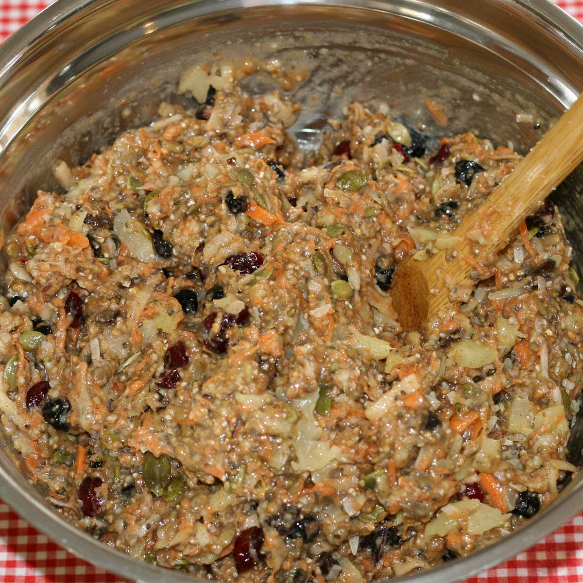 Stainless steel bowl filled with a chunky mixture of muffin batter, grated carrots, seeds, dried fruit, stirred with a wooden spoon.