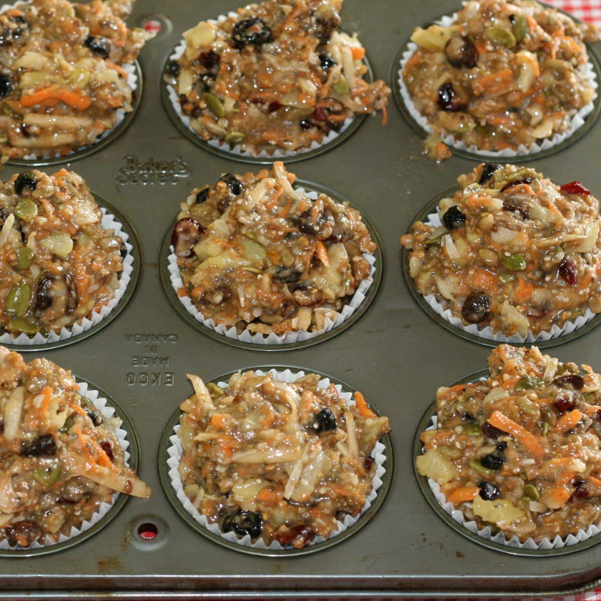 Muffin tin filled with unbaked muffins packed with visible ingredients like nuts, carrots, raisins, and seeds. The mixture looks hearty and rustic.