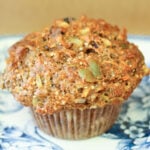Whole grain muffin with nuts and seeds on a blue floral plate. The muffin has a golden-brown top and a rustic, hearty texture.