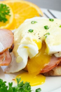 Close-up of Eggs Benedict with Canadian bacon on toasted muffins, topped with Hollandaise sauce, garnished with chopped chives. Orange slices and parsley on the side.