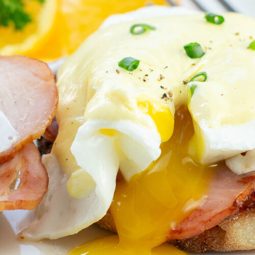 Close-up of Eggs Benedict with Canadian bacon on toasted muffins, topped with Hollandaise sauce, garnished with chopped chives. Orange slices and parsley on the side.