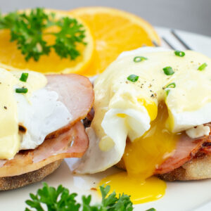 Close-up of Eggs Benedict with Canadian bacon on toasted muffins, topped with Hollandaise sauce, garnished with chopped chives. Orange slices and parsley on the side.
