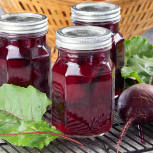Three jars filled with vibrant red pickled beets are placed on a cooling rack next to fresh beetroot and greens
