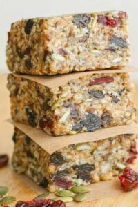 Three stacked fruit-and-seed snack bars, layered with parchment and surrounded by pumpkin seeds and dried red berries on a wooden surface.
