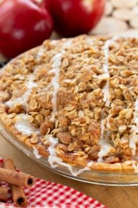 Golden apple crumble pie topped with crumble and white glaze, in a glass dish with red apples, cinnamon sticks and a cozy checkered cloth.