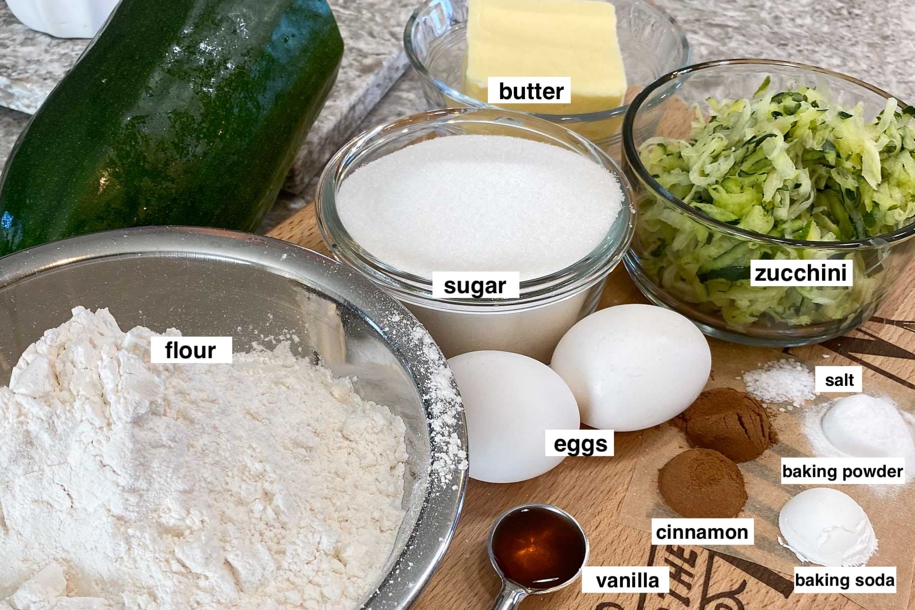 Labeled baking ingredients—flour, sugar, butter, eggs, zucchini, cinnamon, vanilla, salt, baking powder and soda—arranged on a counter.