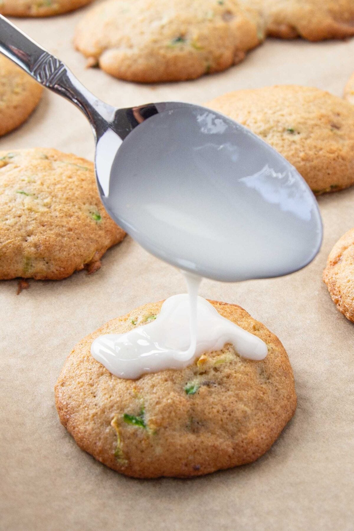 A silver spoon drizzles white icing onto a golden, zucchini cookie, surrounded by freshly baked cookies on parchment paper.