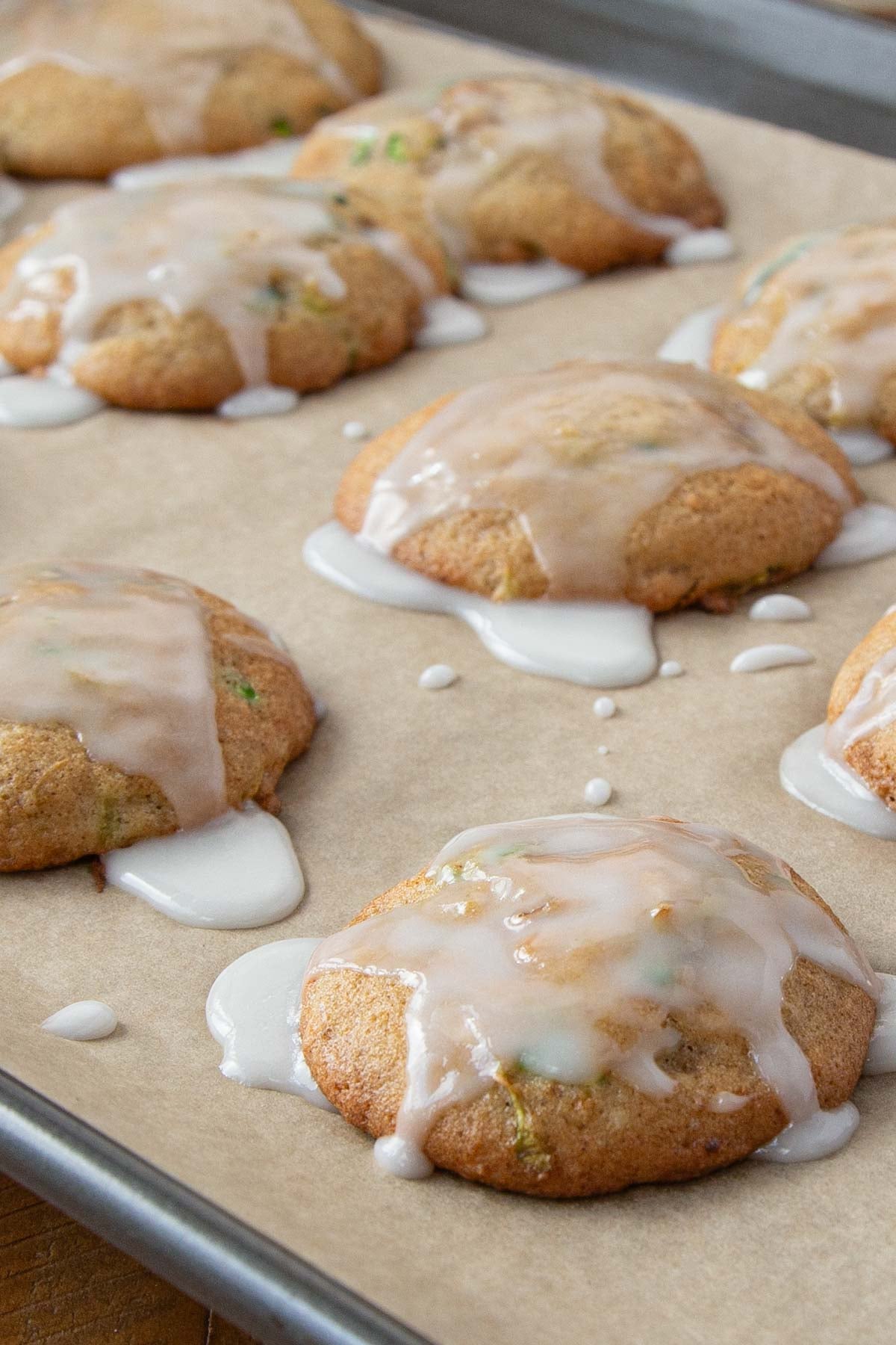 Golden zucchini cookies topped with white glaze dripping over the edges, on a parchment-lined baking sheet.