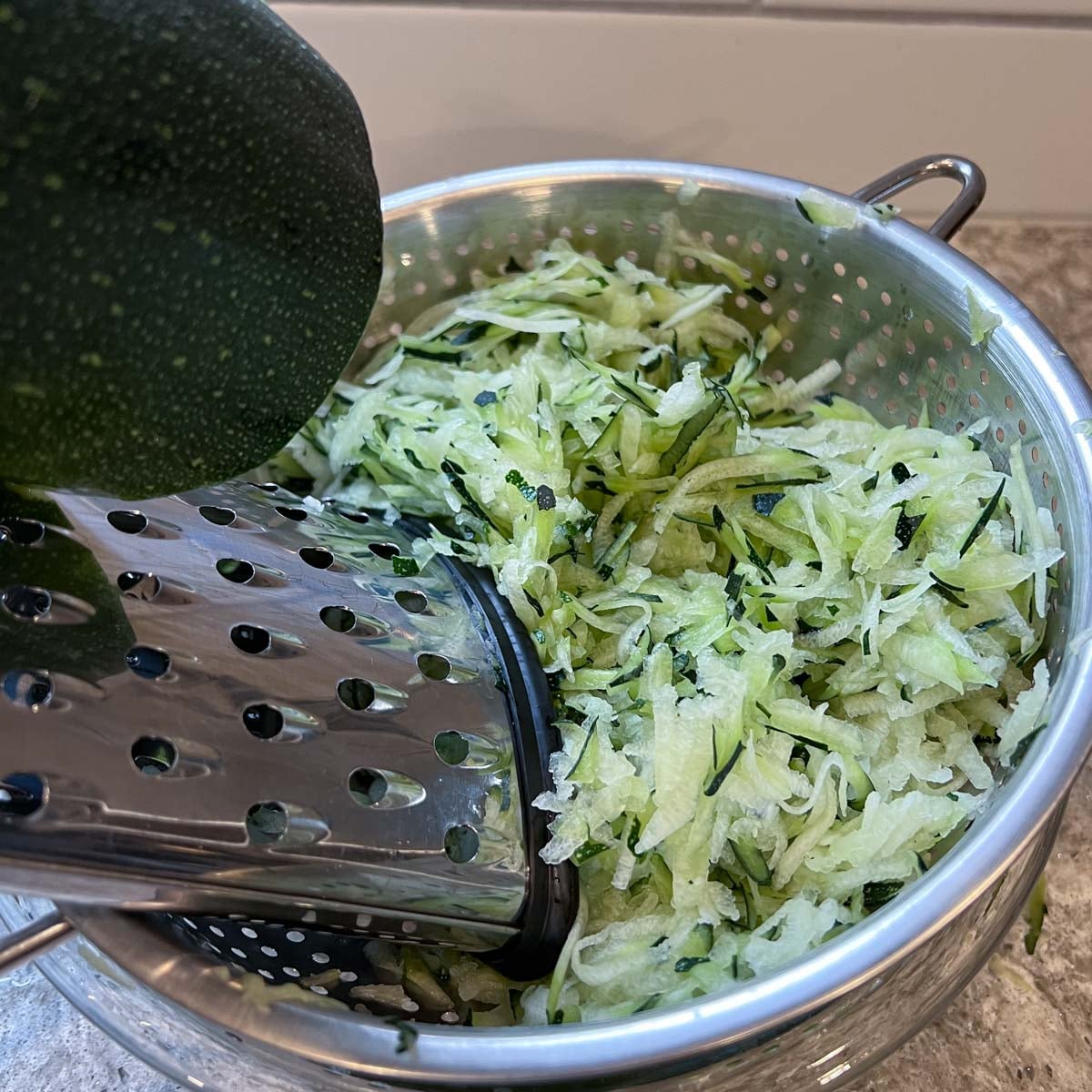 Freshly grated zucchini fills a metal colander beside a box grater and whole zucchini.