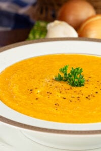 A white bowl of creamy golden-orange butternut squash soup, garnished with fresh parsley and black pepper with onions in the background.